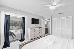 A bedroom with a bed, a dresser, a TV, a ceiling fan, and a view of an outdoor swimming pool.