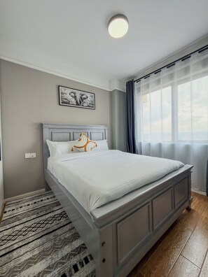 Un dormitorio con cama, luz de techo, ventana con cortinas, un cuadro colgado en la pared y alfombra estampada.