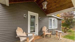 En träveranda med två Adirondack-stolar och ett litet bord, en dörr och ett fönster med utsikt över träd.