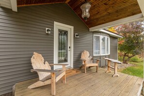 Eine Holzterrasse mit zwei Adirondack-Stühlen und einem kleinen Tisch, einer Tür und einem Fenster mit Blick auf Bäume.