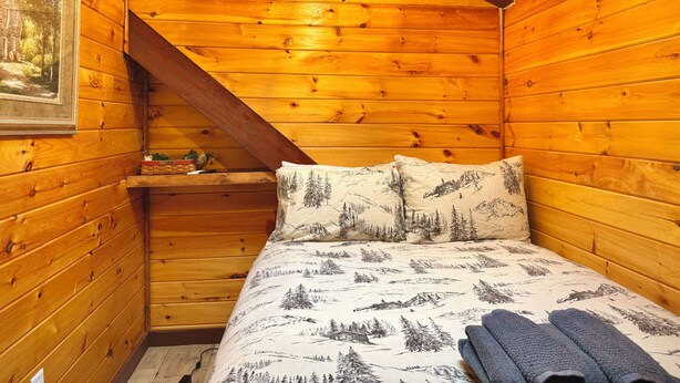 A wooden cabin room with a bed, a painting on the wall, and a small shelf with items.