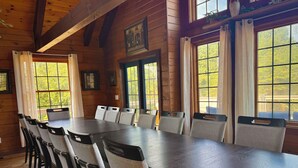 A long wooden dining table with multiple chairs, large windows with curtains, and framed pictures on the walls.