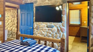 A bedroom with a stone wall, a blue door, a flat-screen TV mounted on the wall, a bed with a checkered bedspread, and a wooden bench. - Luxury Renovated | Pool | Hot Tub | Sleeps 20 (New Plymouth)