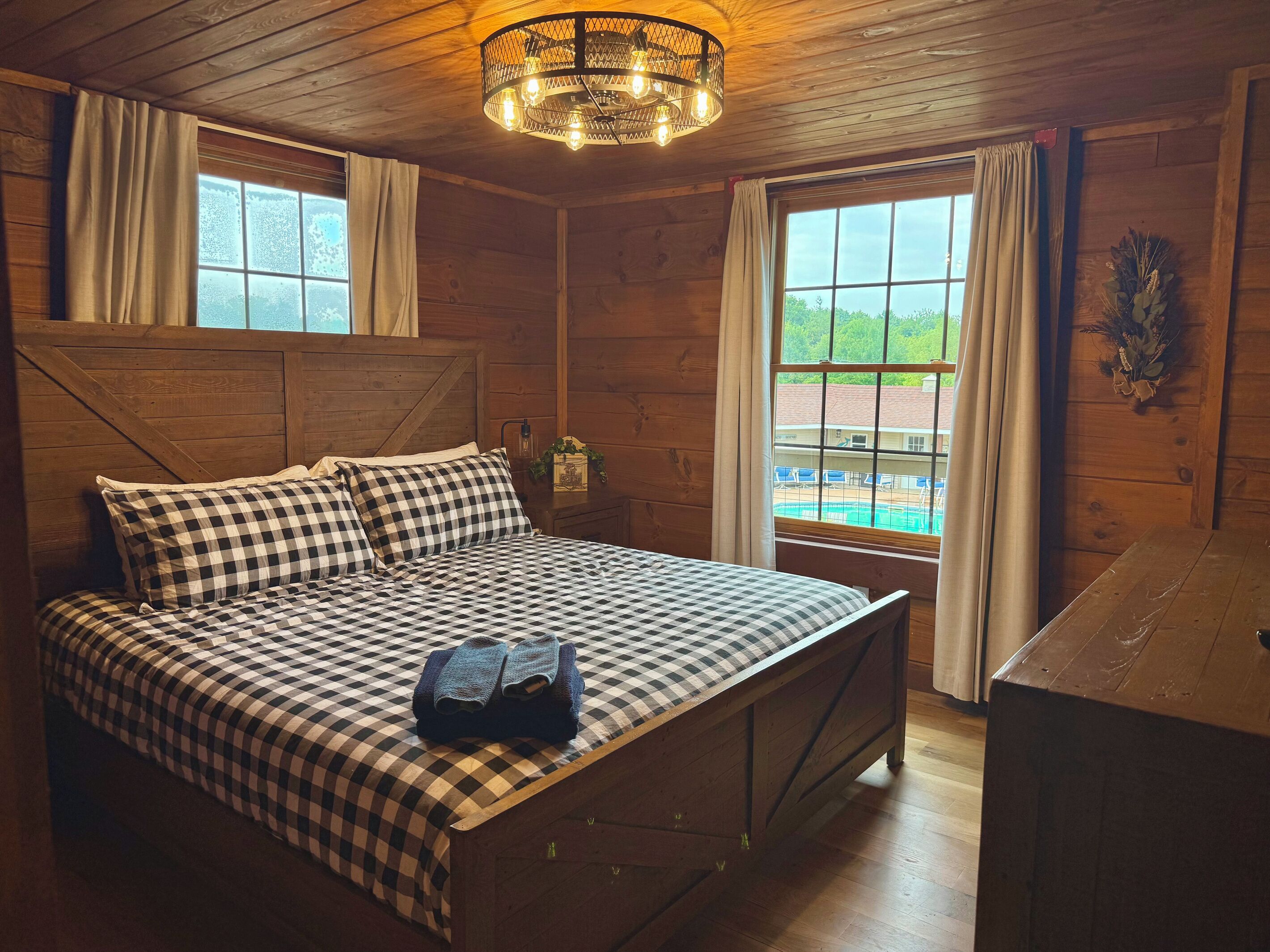 A wooden cabin room with a bed, a desk, and a window overlooking a pool.