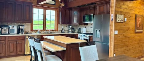 Una cocina con armarios de madera, un refrigerador de acero inoxidable, una isla grande con taburetes de bar y una ventana con vistas a un paisaje verde.