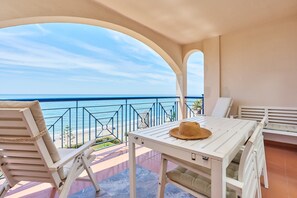 Ein Balkon mit einem weißen Tisch und Stühlen, ein Strohhut auf dem Tisch und Blick aufs Meer.