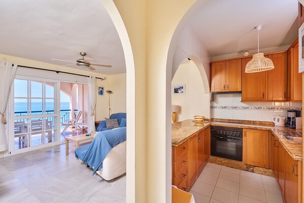 A room with a bed, a balcony with a view, a kitchen with wooden cabinets, and a ceiling fan. - Apartment 'Blue View' with Sea View, Wi-Fi and Air Conditioning (Torrox Costa)