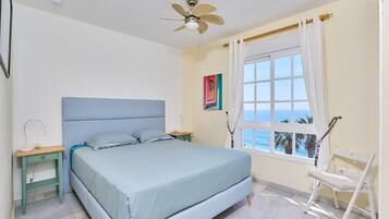 A bedroom with a bed, a ceiling fan, a nightstand, a chair, a window with curtains, and a painting on the wall.