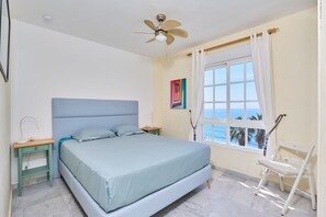 Ein Schlafzimmer mit Bett, Deckenventilator, Nachttisch, Stuhl, Fenster mit Vorhängen und einem Bild an der Wand.