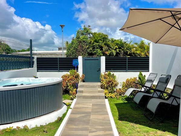 A modern outdoor patio with a hot tub, lounge chairs, and a pathway leading to a gated entrance. - Vrbo Property (Les Abymes)
