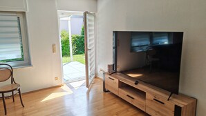Habitació amb un televisor de pantalla gran muntat sobre un suport de fusta, una cadira de vimetla i una porta corredissa de vidre que dona a una zona exterior.