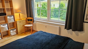 A room with a bed, a desk, a chair, a radiator, and a window with curtains.