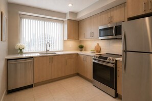 A modern kitchen with stainless steel appliances, wooden cabinets, and a window with blinds. - The Elowyn Hotel (Toronto)