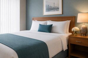 A neatly made bed with white and teal bedding, a wooden headboard, a nightstand with a lamp and flowers, and a framed cityscape picture on the wall. - The Elowyn Hotel (Toronto)