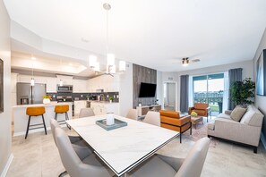 Eine moderne Küche mit einer zentralen Kochinsel, ein Essbereich mit einem Marmortisch und ein Wohnbereich mit einer Couch und Fernseher.