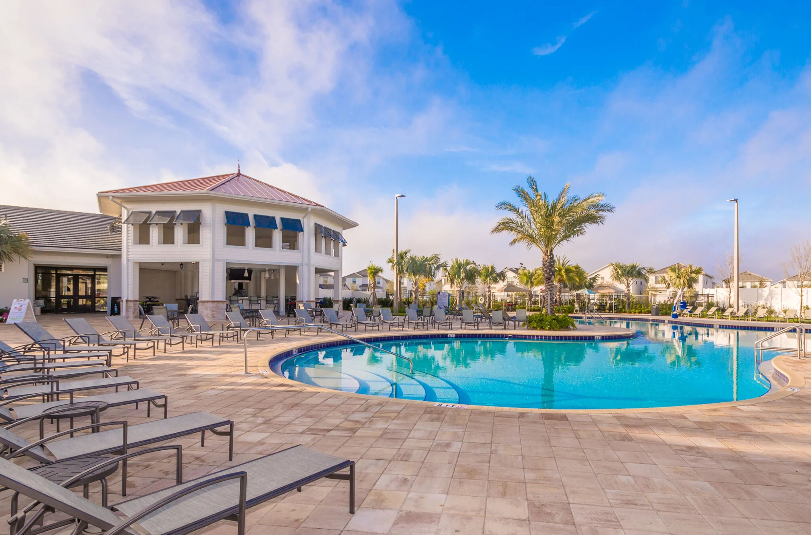 A pool area with lounge chairs, a building with a red roof, and palm trees.