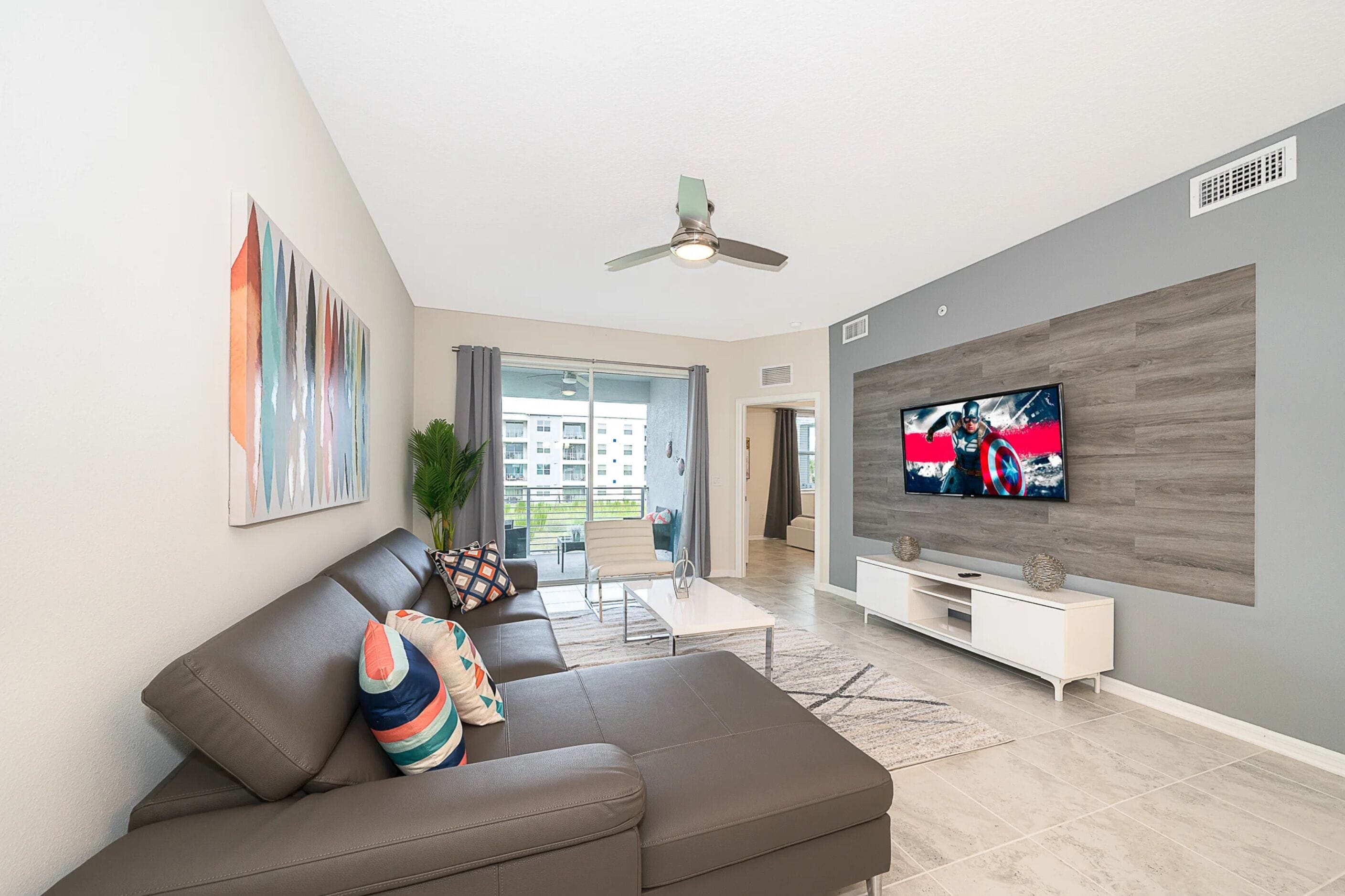A modern living room with a sectional sofa, a flat-screen TV, a ceiling fan, and a large window.