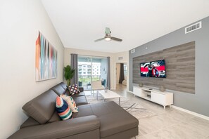 Ein modernes Wohnzimmer mit einem Ecksofa, einem Flachbildfernseher, einem Deckenventilator und einem großen Fenster.