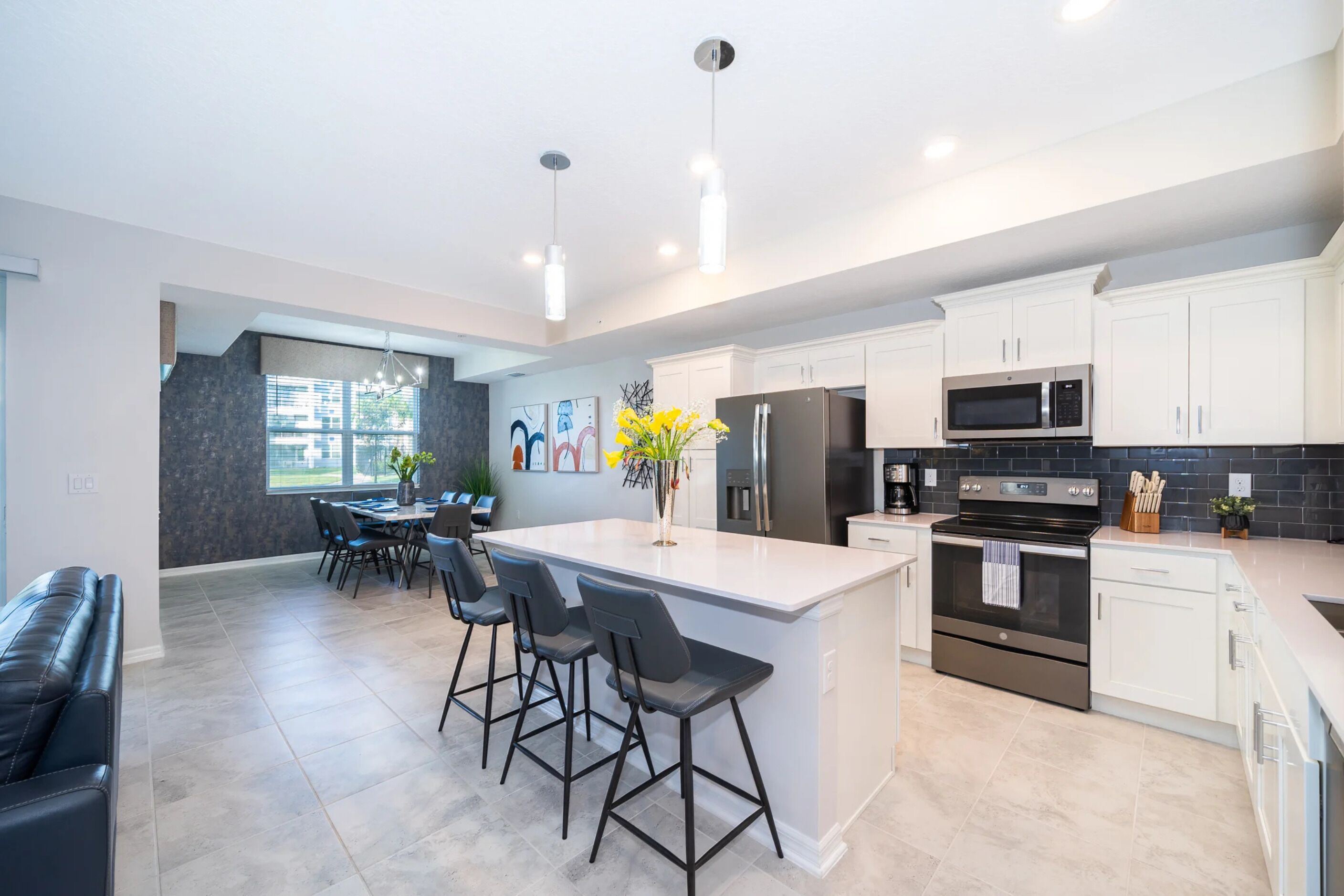 A modern kitchen with white cabinets, a central island with bar stools, stainless steel appliances, and a dining area with a table and chairs.