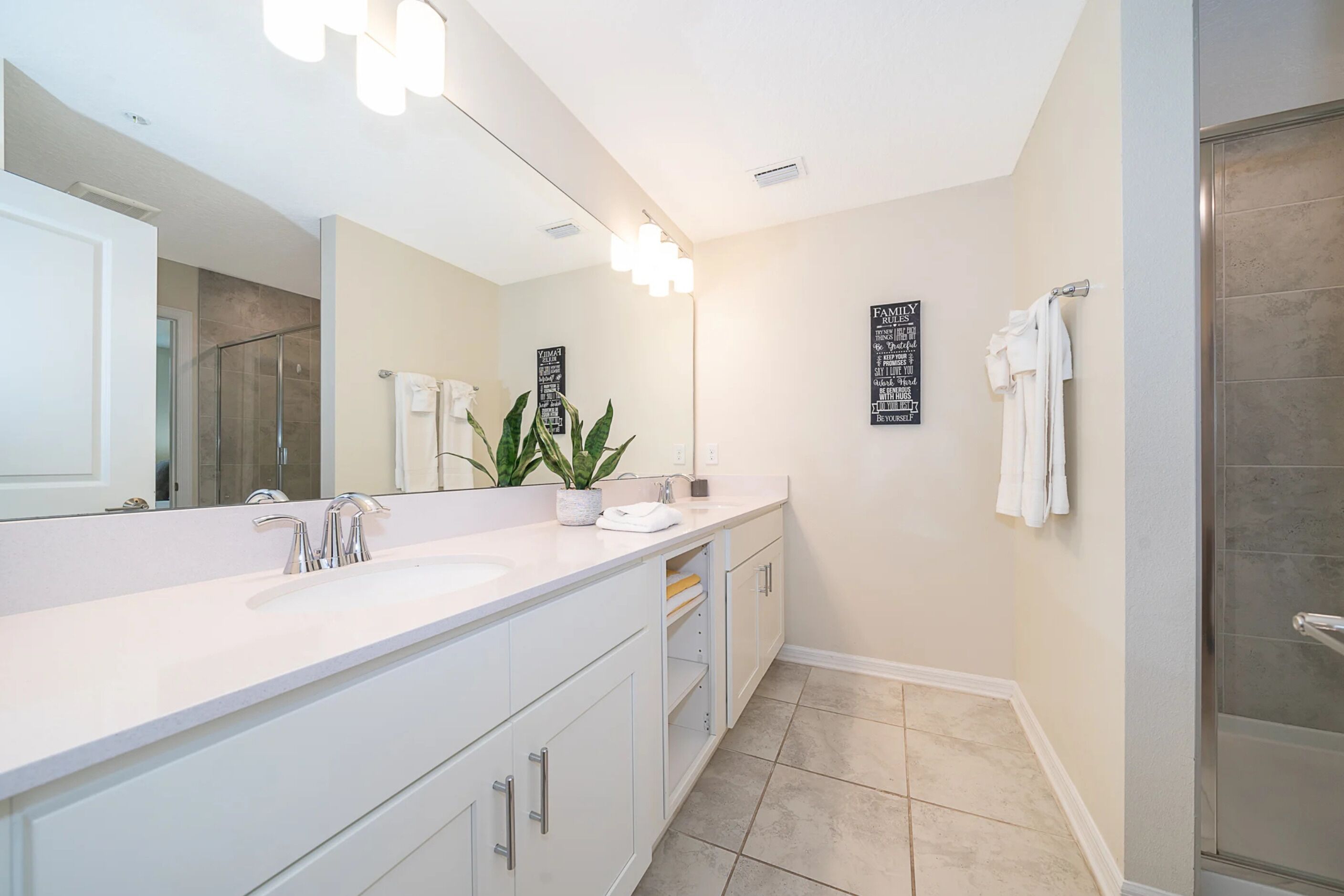 A modern bathroom with a large mirror, two sinks, a shower, and a potted plant.