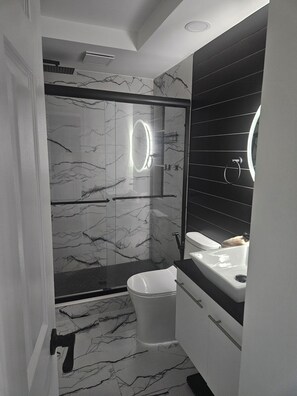 Et moderne badeværelse med brusekabine i glas, marmorvægge, et hvidt toilet og et sort toiletbord.