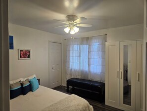 A bedroom with a bed, a ceiling fan, a bench, a wardrobe, and a window with sheer curtains.
