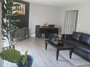 En stue med en sort sofa, et sofabord, en potteplante, en pejs og et billede på væggen.