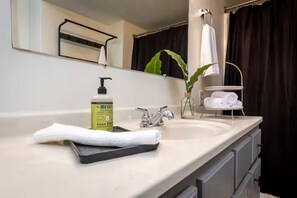Une salle de bain avec un lavabo, un miroir, un porte-serviettes, une plante et un flacon de savon pour les mains.