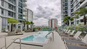 Una piscina en la azotea con sillones de descanso, palmeras y edificios modernos al fondo.
