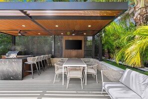 Eine Terrasse im Freien mit Esstisch, Stühlen, einer Couch, einem Fernseher und einem Deckenventilator.