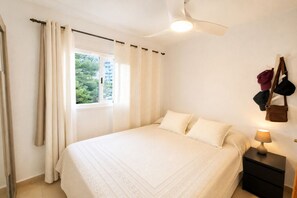 Ein Schlafzimmer mit Bett, Vorhängen, einem Nachttisch mit Lampe und Blick auf Grünflächen.