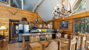 L’intérieur d’une cabane avec des murs en bois, un coin repas, une cuisine équipée et une grande fenêtre donnant sur une forêt.