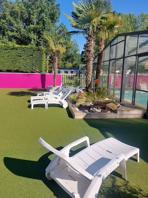 Een zwembadgedeelte met kunstgras, witte lounge fauteuils, palmbomen en een glazen constructie.