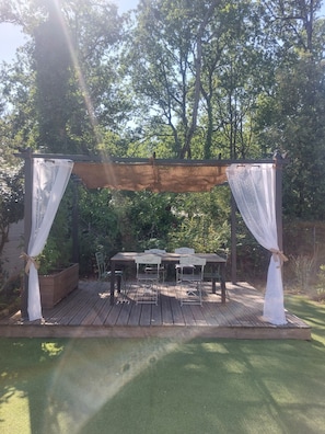 Een terras van hout buiten, met een pergola, witte gordijnen en een tafel met stoelen.