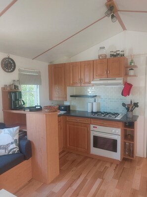 A compact kitchen with wooden cabinets, a gas stove, a built-in oven, and a window with a striped valance. - Enjoy a relaxing vacation in this 4-person mobile home (notre dame de riez)