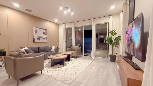 A modern living room with a sofa, coffee table, TV, and a sliding glass door leading to a balcony. - 4BR Private Pool Home with Lake View Near Disney | Sleeps 10 (Clermont)