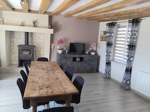 Een eetruimte met een houten tafel en stoelen, een stenen open haard, een televisie op een kast en een raam met gordijnen met een patroon.