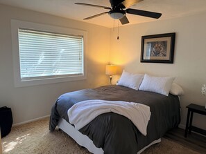 Une chambre à coucher avec un lit, un ventilateur de plafond, une fenêtre avec des stores, une lampe et un tableau encadré au mur.