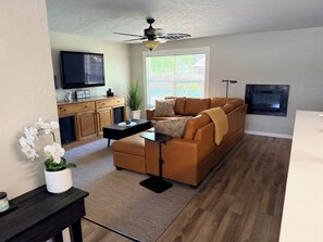 Un salon comprenant un canapé, une table basse, un tél éviseur, un ventilateur de plafond, une fenêtre avec des stores et une cheminée.