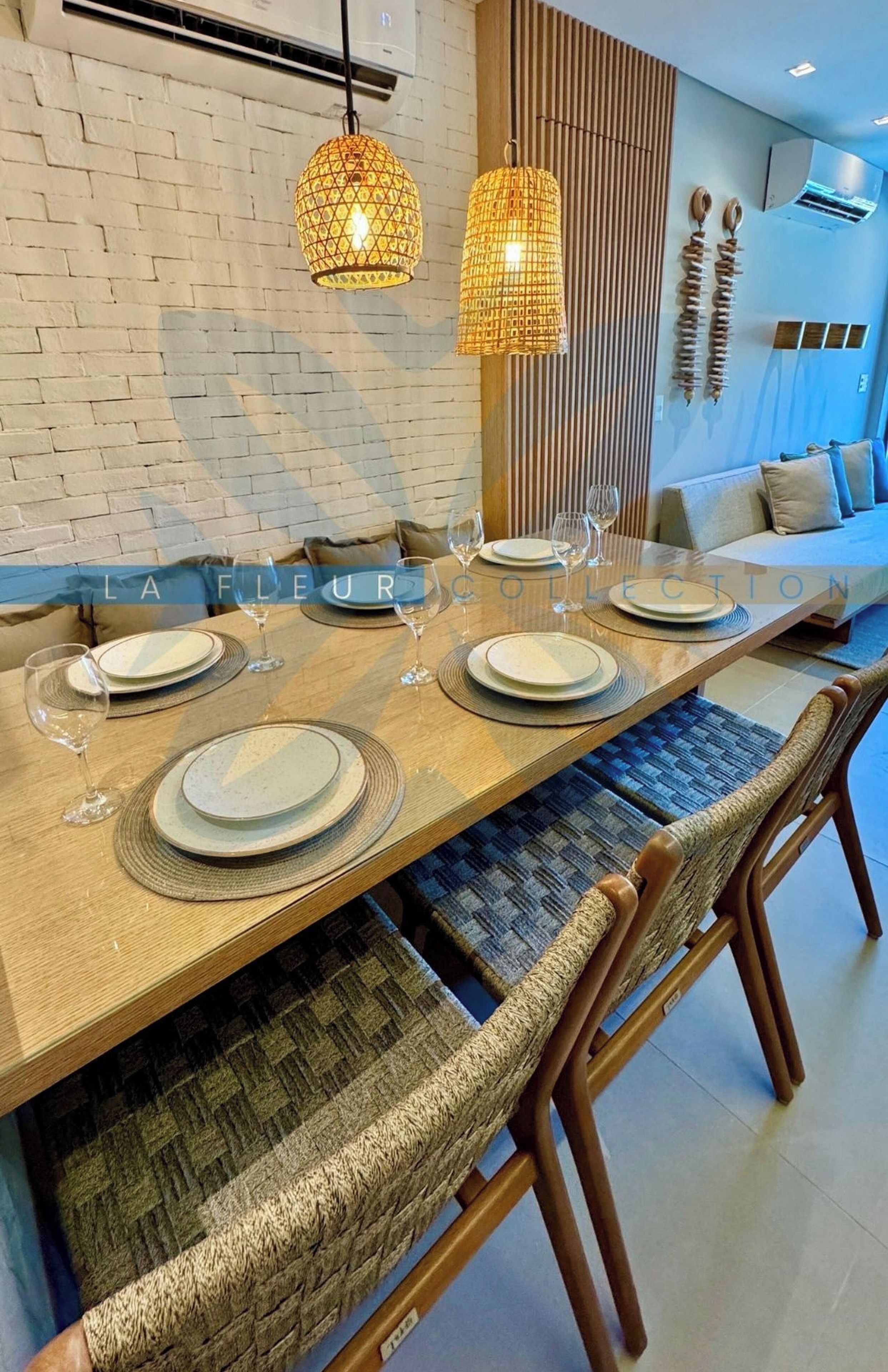 A dining area with a long table set for four, surrounded by woven chairs, with a brick wall and wooden slats in the background.
