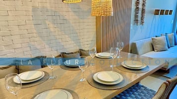 A dining area with a long table set for four, surrounded by woven chairs, with a brick wall and wooden slats in the background.