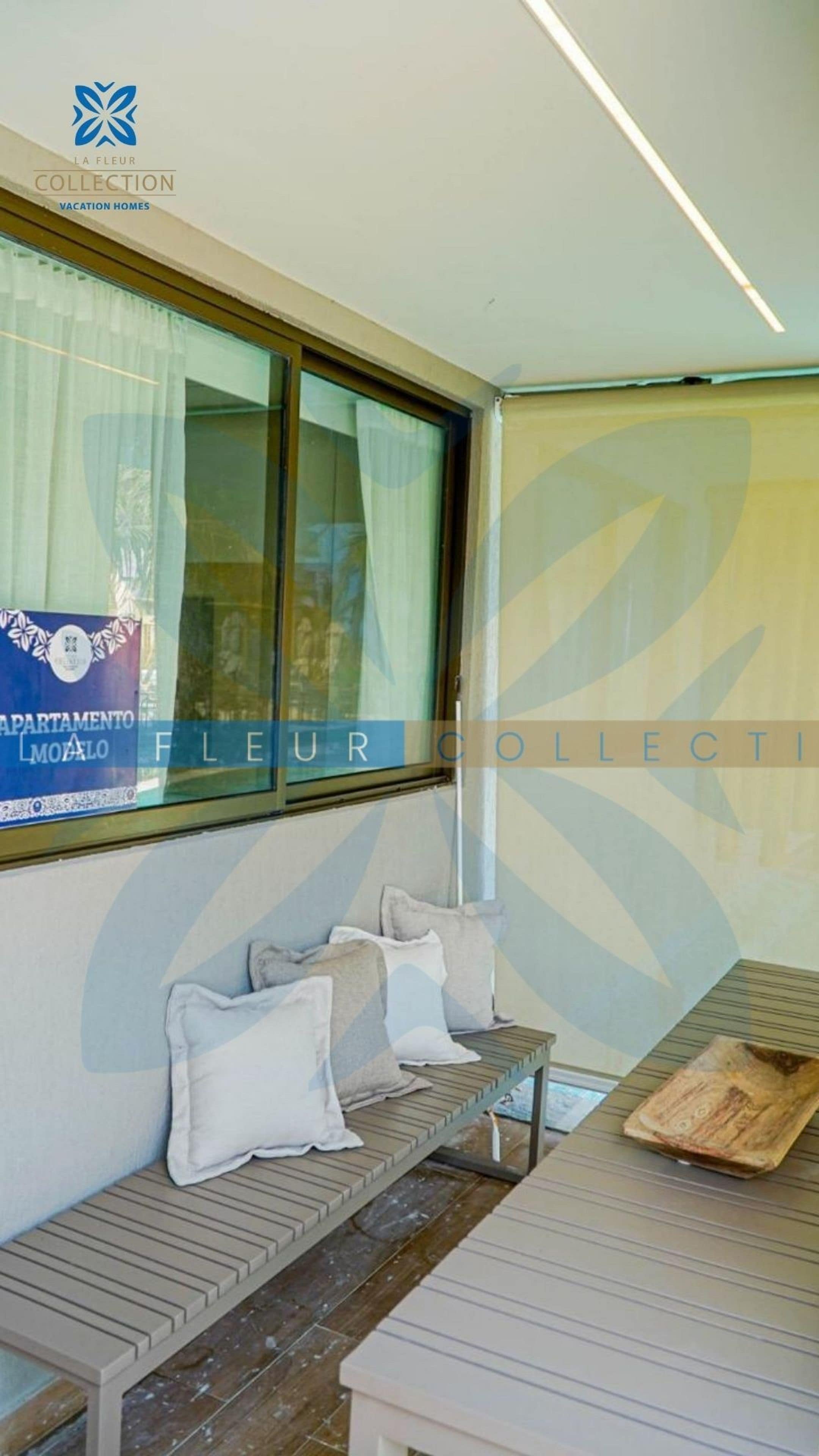 A room with a bench, a table, and a window with curtains. There is a sign on the window and a logo on the wall.