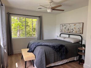A bedroom with a bed, a bench, a ceiling fan, a window with curtains, a wall-mounted artwork, and a bedside table with a plant.