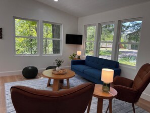 A living room with a blue sofa, two brown chairs, a round wooden table, and a TV mounted on the wall.