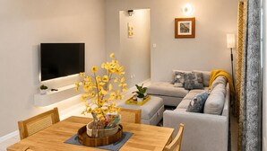 Uma sala de estar moderna com televisão de tela plana, uma mesa de jantar de madeira e um sofá com almofadas estampadas.