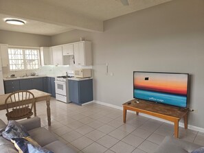Ett modernt kök med vita skåp, en mikrovågsugn och en spis. Här finns också en matplats med ett bord och stolar, och en tv-apparat på ett stativ som visar en solnedgång.