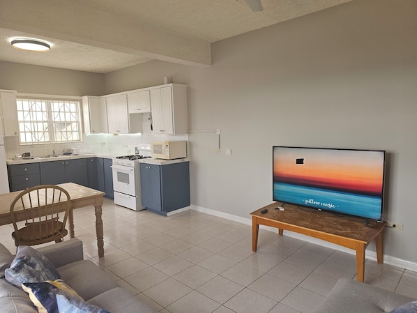 Eine moderne Küche mit weißen Schränken, einer Mikrowelle und einem Herd. Es gibt einen Essbereich mit Tisch und Stühlen, und ein Fernseher auf einem Schrank, der eine Sonnenuntergangszene zeigt.