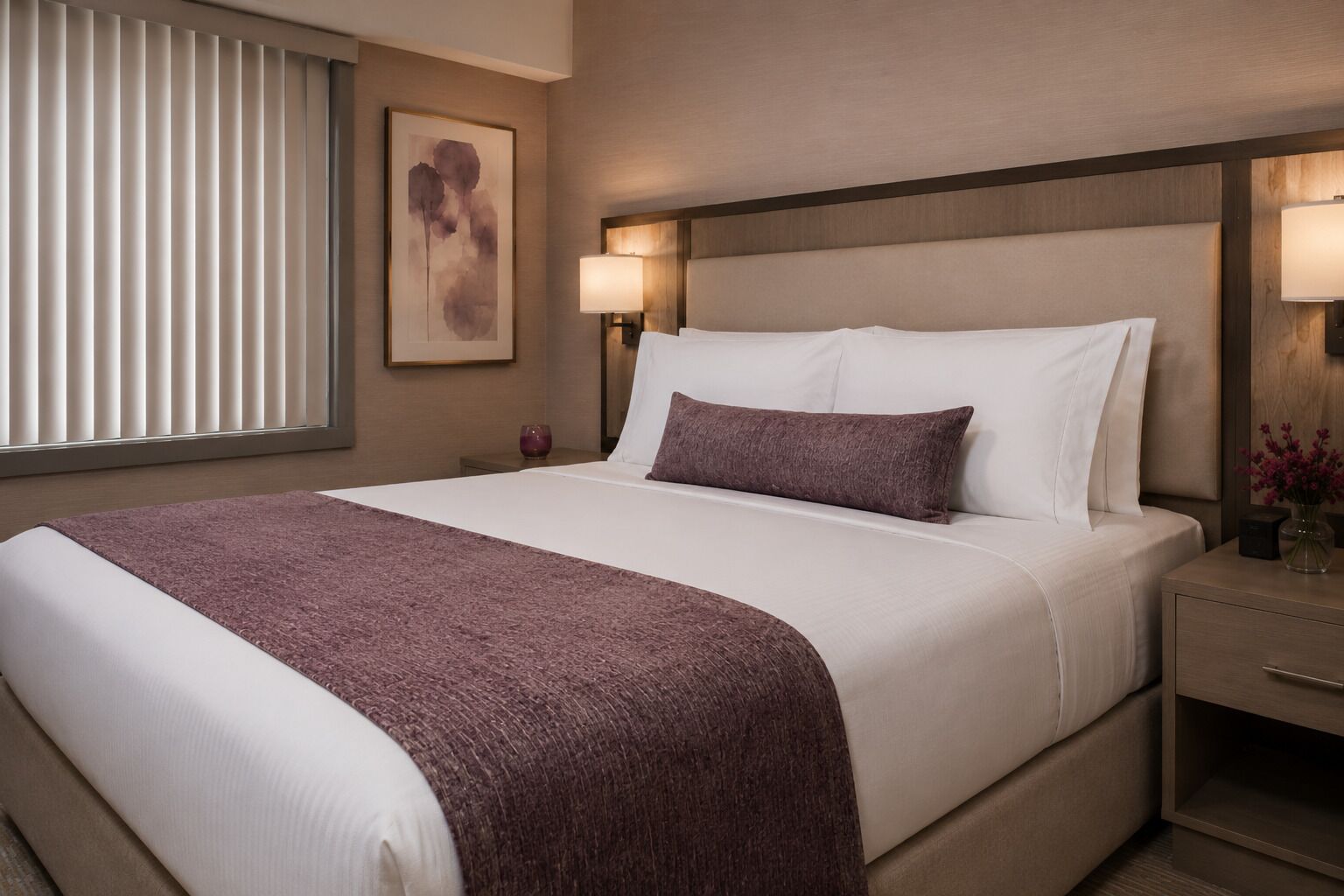 A hotel room with a bed, bedside lamps, a nightstand, a framed picture on the wall, and a window with blinds.