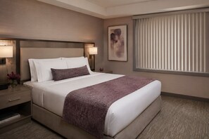 A hotel room with a bed, bedside lamps, a nightstand, a framed picture, and a window with blinds. - The Ravona Suites (Toronto)