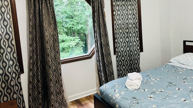 Une chambre avec une fenêtre, des rideaux, un lit recouvert d’une couvre-lit à motifs floraux et une tête de lit en bois.